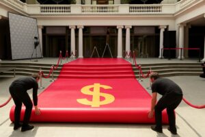 Two workers unrolling a red carpet with a large yellow dollar sign at the center.
