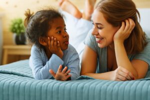 A smiling mother and her young daughter conversing on the bed, smiling.