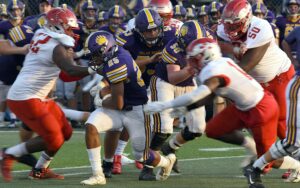 A high school football running back in a purple uniform carries the ball as teammates block defenders in red.
