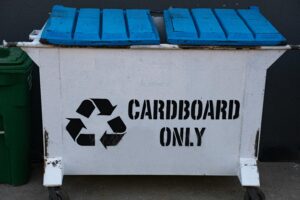 White recycling dumpster labeled "Cardboard Only" with blue lids and a recycling symbol.